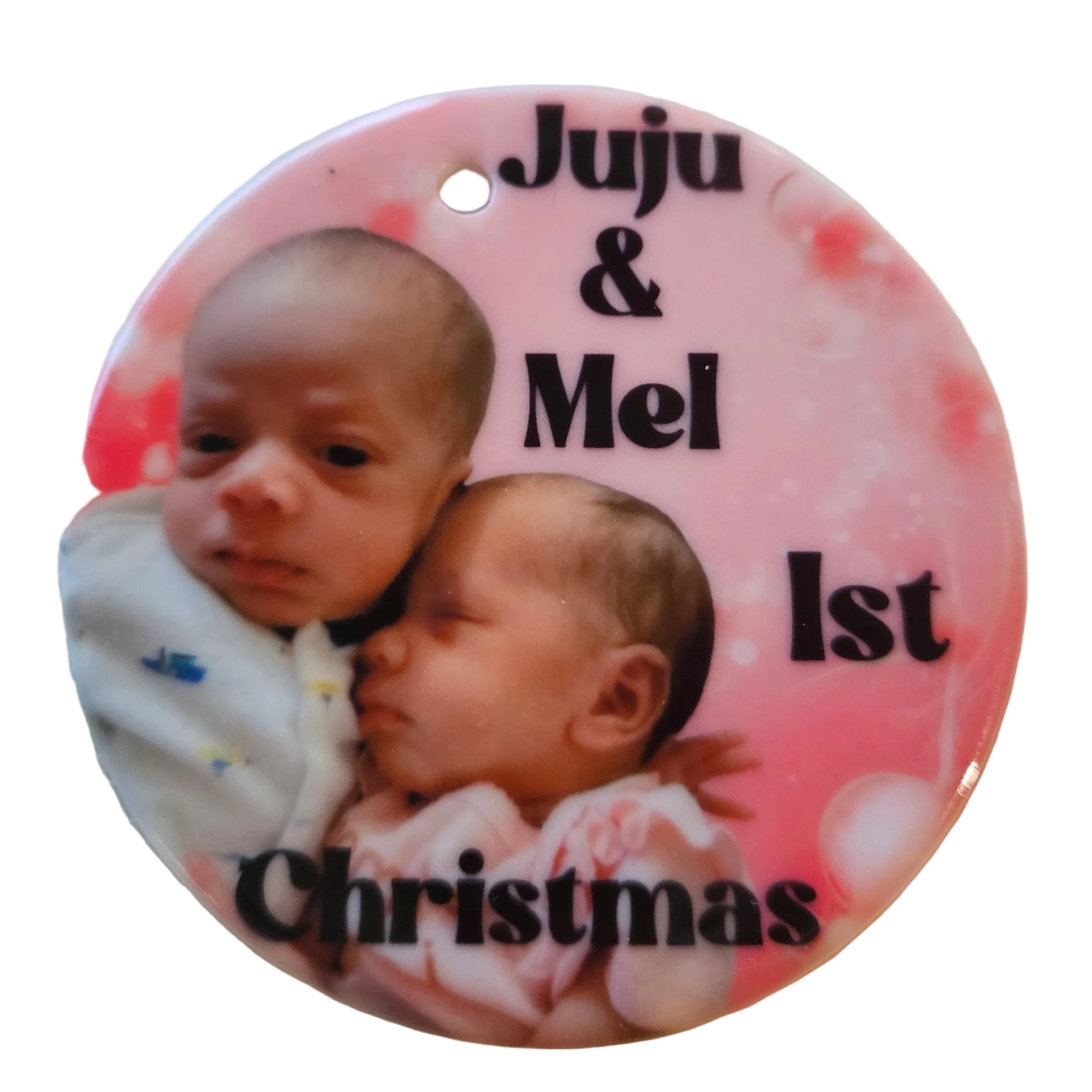 Round pink ornament with two babies and text 'Juju & Mel 1st Christmas' on a textured surface.